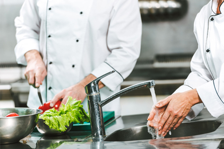 Chefs in a commercial kitchen — one cutting vegetables, one washing hands at a sink